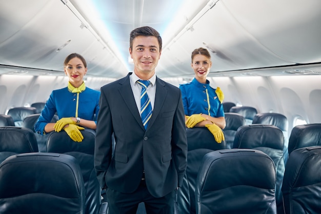 Ritratto in vita di un bel assistente di volo maschio con le sue due colleghe in uniforme elegante in piedi sul bordo dell'aereo passeggeri