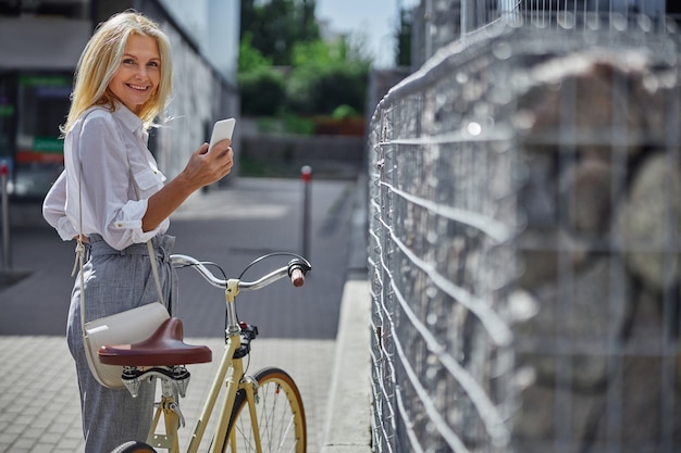 Ritratto in vita di una donna allegra felice in camicetta bianca che tiene il telefono cellulare mentre si cammina con la bici in città