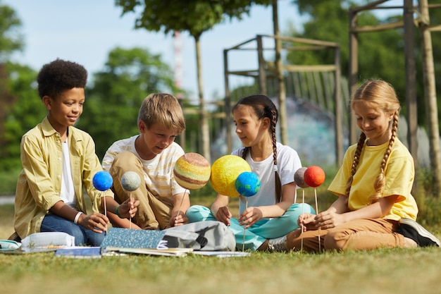 Ritratto integrale dei bambini multietnici del gruppo o che si siedono sull'erba verde e che tengono i pianeti modello mentre godono della lezione di astronomia all'aperto alla luce del sole