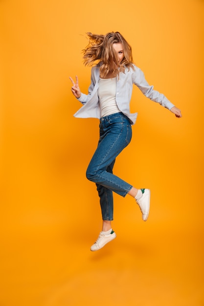 Ritratto integrale dei jeans e delle scarpe da tennis d'uso della donna allegra che saltano o che corrono con il gesto di vittoria, isolato sopra fondo giallo