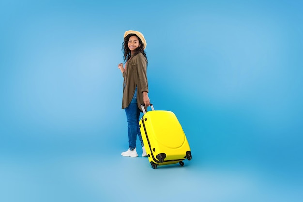 Ritratto integrale del viaggiatore femminile nero felice che cammina con la valigia sopra lo studio blu