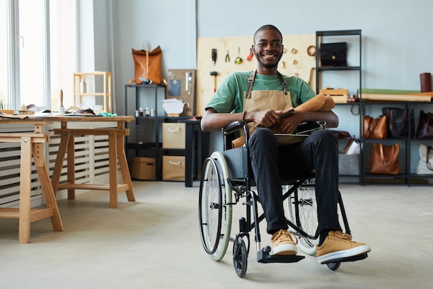 Ritratto integrale dell'uomo afroamericano sorridente che utilizza la sedia a rotelle che si gode il lavoro in pellettieri...