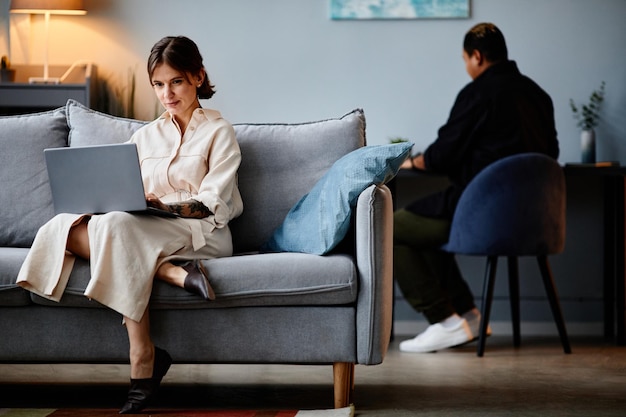 Ritratto integrale della giovane donna moderna che si rilassa sul divano a casa e utilizza lo spazio della copia del computer portatile