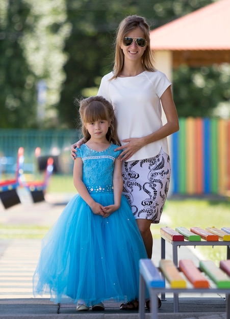 Ritratto integrale della giovane madre sorridente sottile bionda felice in occhiali da sole e piccola ragazza graziosa della figlia in vestito da sera blu lungo sull'asilo offuscato parco giochi.