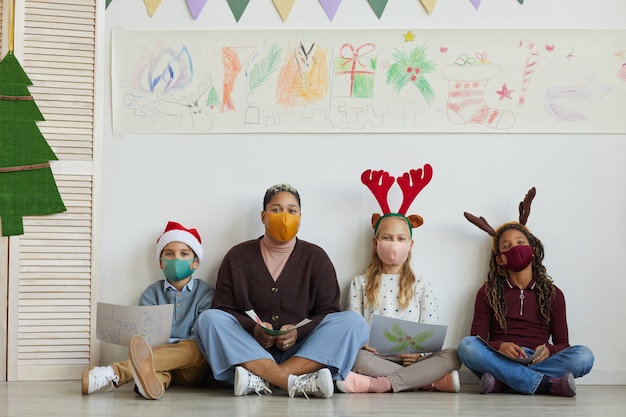 Ritratto integrale della maschera da portare dell'insegnante femminile mentre era seduto sul pavimento con un gruppo multietnico di bambini che tengono le immagini durante la lezione di arte a Natale, lo spazio della copia