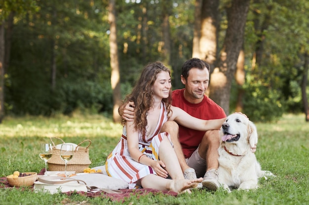 Ritratto integrale di giovane coppia con il cane che gode del picnic all'aperto sullo spazio della copia dell'erba verde