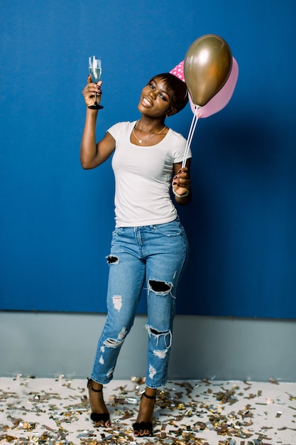 Ritratto integrale di giovane donna africana allegra che tiene mazzo di aerostati e di champagne isolati sopra spazio blu. Festa di compleanno, concetto di celebrazione.