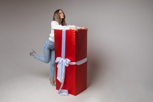 Ritratto integrale di giovane donna attraente con capelli biondi in maglione bianco casual, jeans e tacchi che abbracciano grande regalo avvolto rosso con fiocco bianco con labbra imbronciate