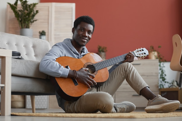 Ritratto integrale di giovane uomo afro-americano a suonare la chitarra e sorridere alla telecamera mentre era seduto sul pavimento a casa, copia dello spazio