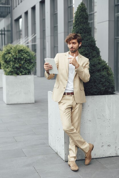 Ritratto integrale di giovane uomo d'affari barbuto in vestito beige che ha videochiamata sulla tavoletta digitale all'aperto