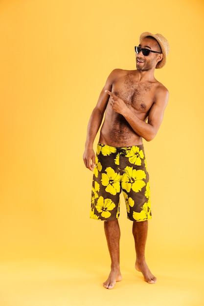 Ritratto integrale di un uomo afro sorridente in costume da bagno che punta il dito lontano isolato su una parete arancione