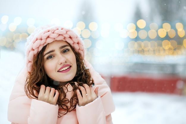 Ritratto invernale all'aperto di una ragazza sorridente attraente che indossa cappotto rosa e berretto lavorato a maglia in posa vicino alle luci. Spazio per il testo