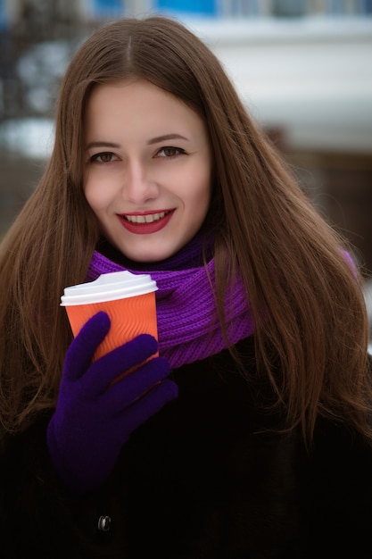 Ritratto invernale di allegra donna bruna che indossa una sciarpa lavorata a maglia viola e tiene in mano una tazza di tè