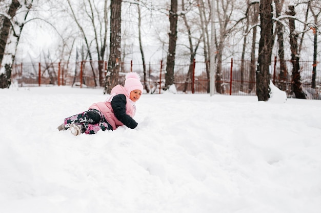 Ritratto invernale di bambina sorridente e sdraiata fuori nella neve in abiti invernali caldi con alberi ad alto fusto intorno Adorabile sfondo pieno di neve