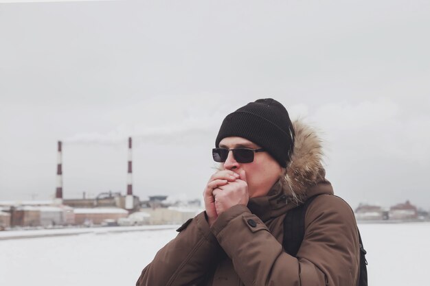 Ritratto invernale di giovane uomo in abiti invernali casual con occhiali sulla città industriale dell'argine a piedi