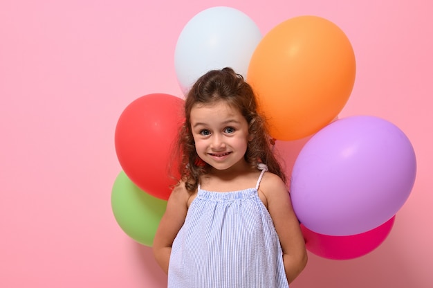 Ritratto isolato, colpo alla testa, primo piano di una bellissima bambina allegra di 4 anni con palloncini multicolori che sorride guardando la fotocamera. Sfondo rosa, spazio per il testo, fotografia pubblicitaria