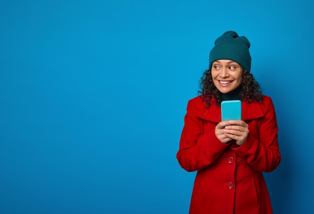 Ritratto isolato su sfondo blu di un allegro fantastico felice bella donna che indossa un cappotto rosso brillante e un caldo cappello di lana verde che tiene il telefono cellulare e si rallegra guardando da parte. Copia spazio pubblicitario