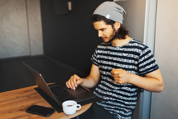 Ritratto laterale di giovane uomo bello che lavora a casa sul computer portatile; tazza di caffè e smartphone sul tavolo di legno.