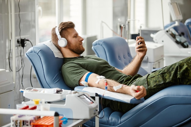 Ritratto laterale di un giovane sorridente che dona sangue in tutta comodità e guarda video tramite smartphone