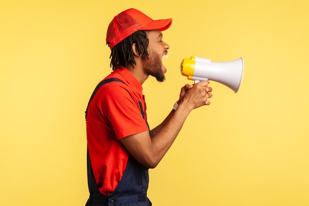 Ritratto laterale di un lavoratore aggressivo che indossa una maglietta rossa, un berretto e una tuta blu che tiene il megafono e urla forte, protestando. Studio indoor girato isolato su sfondo giallo.