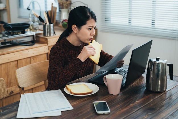 ritratto laterale infastidita casalinga asiatica che mangia colazione sta tenendo e fissando il documento di legge con un'espressione seria mentre presenta la tassa sul computer a casa.