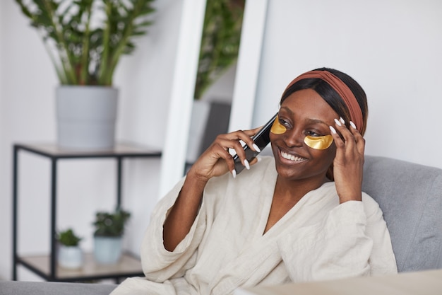 Ritratto minimo di bella donna afro-americana che parla tramite smartphone mentre si gode la routine di cura della pelle a casa, copia spazio