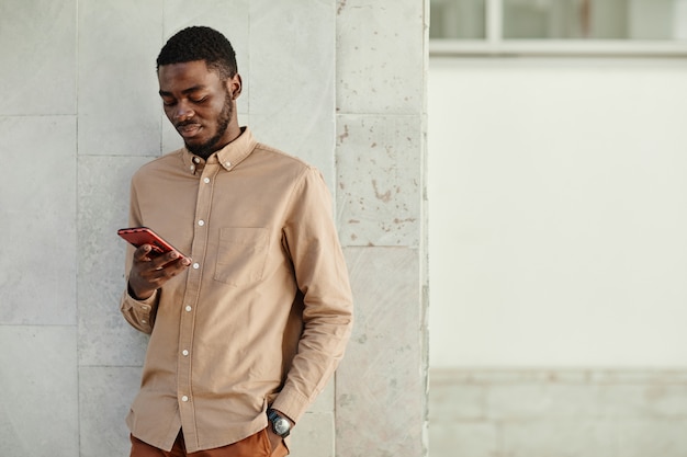 Ritratto minimo di giovane uomo afro-americano che utilizza lo smartphone mentre si trova sullo sfondo urbano, copia spazio