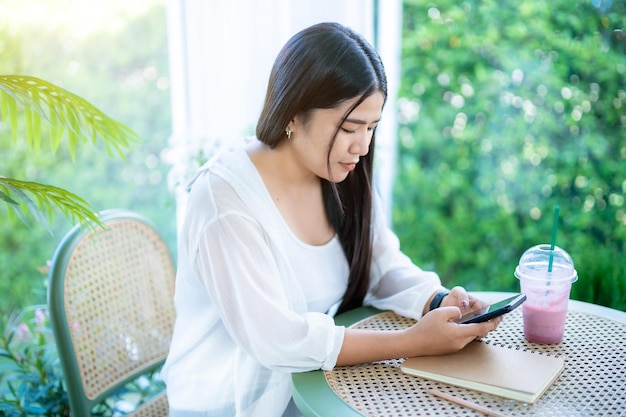 Ritratto moderno di donna asiatica felice che indossa smartwatch che indossa un bel vestito bianco e indossa jeans stile di vita godendo di parlare facendo utilizzando smartphone in seduta caffetteria ristorante sfondo