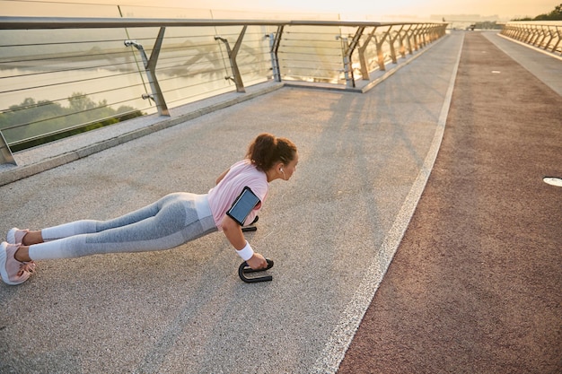 Ritratto orizzontale a figura intera di una sportiva, atleta femminile che fa push-up mentre pratica un allenamento incrociato, allenamento a corpo libero sul ponte della città all'alba. Sport, concetto di stile di vita attivo