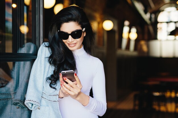 Ritratto orizzontale della donna magnifica del brunette in vestiti bianchi ed occhiali da sole che si siedono in caffè facendo uso del suo smartphone