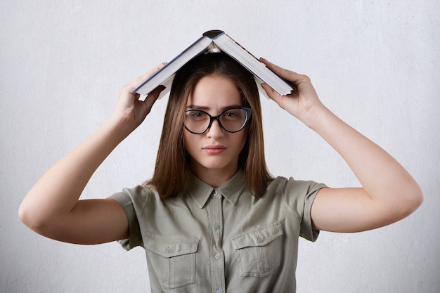 Ritratto orizzontale della giovane studentessa indossando occhiali e camicia tenendo il libro sulla testa essendo stanco di studiare.