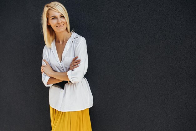 Ritratto orizzontale di bella giovane donna bionda sorridente in piedi con le braccia incrociate e guardando la fotocamera contro il muro grigio sulla strada Ritratto schietto di donna d'affari Spazio di copia