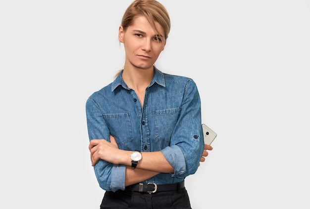 Ritratto orizzontale interno di una donna bionda seria freelance con i capelli biondi vestita con una camicia di jeans blu in posa sullo sfondo dello studio Donna d'affari con il telefono cellulare in posa sul muro bianco