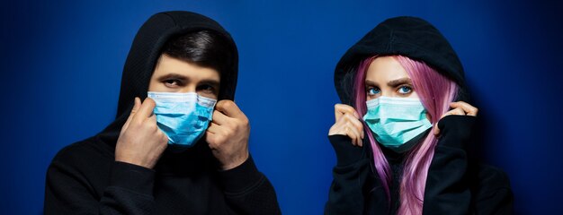 Ritratto panoramico in studio scuro di giovane coppia incappucciata, ragazzo e ragazza che indossa una maschera medica contro il coronavirus, su sfondo di colore blu fantasma.