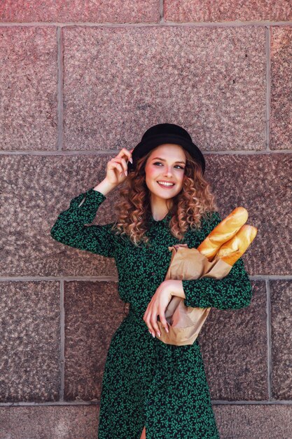 Ritratto piuttosto giovane donna con baguette in mano sulla trama di sfondo della parete. La ragazza vestita in stile francese mostra emozione con il pane. Femmina in vestiti alla moda che tengono baguette fresche. Copia spazio