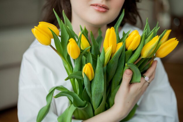 Ritratto primaverile di giovane bella donna con occhi azzurri e capelli ricci con bouquet di tulipani gialli all'interno della casa Modello felice in jeans e camicia bianca Festa della mamma