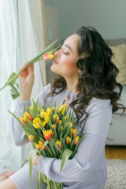 Ritratto primaverile di una bella donna riccia dai capelli scuri in abito lilla con un grande mazzo di tulipani colorati a casa Il modello emotivo tiene i fiori nelle sue mani Festa della donna Presente Festa della mamma