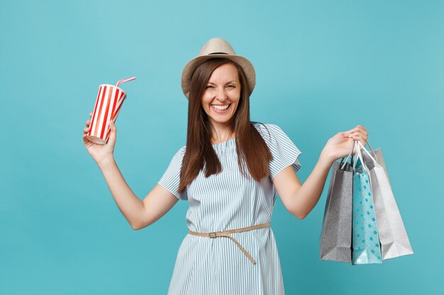 Ritratto ragazza sorridente abbastanza alla moda in abito estivo, cappello di paglia che tiene pacchetti borse con acquisti dopo lo shopping, tazza di acqua gassata isolata su sfondo blu pastello. Copia spazio per la pubblicità