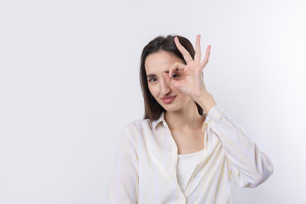 Ritratto ravvicinato di attraente giovane donna eccentrica che fa il binocolo con le mani che mostrano il gesto giusto su sfondo bianco per studio