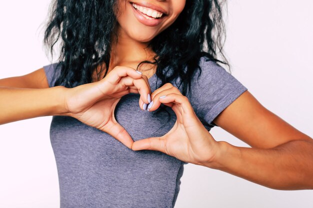 Ritratto ravvicinato di contenuto Ragazza etnica afro con capelli neri corvini disordinati in piedi sorridente ampiamente, con i palmi vicino al petto che mostra un cuore a mano