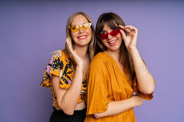 Ritratto ravvicinato di due donne alla moda in occhiali da sole e abiti estivi alla moda in posa su bakground viola in studio
