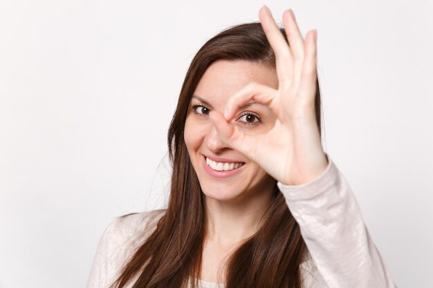 Ritratto ravvicinato di giovane donna sorridente in abiti leggeri che guarda la fotocamera che mostra il gesto OK isolato su sfondo bianco della parete in studio. Persone sincere emozioni, concetto di stile di vita. Mock up spazio di copia.