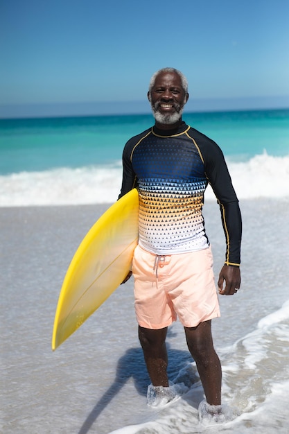 Ritratto ravvicinato di un anziano afroamericano su una spiaggia al sole, con una tavola da surf sotto il braccio, guardando la telecamera e sorridendo, con il cielo blu e il mare sullo sfondo