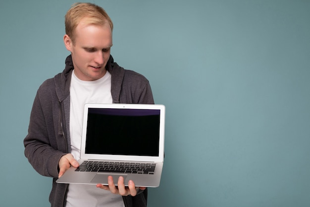 Ritratto ravvicinato di un bell'uomo biondo che tiene in mano un computer portatile che guarda la tastiera del netbook isolato su sfondo blu.