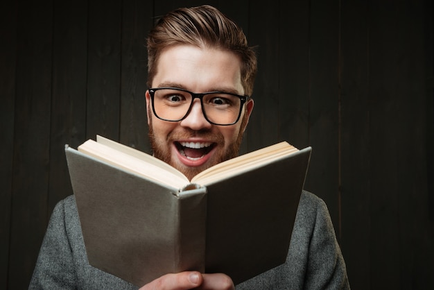 Ritratto ravvicinato di un uomo allegro felice in occhiali che legge un libro isolato sullo sfondo di legno nero black