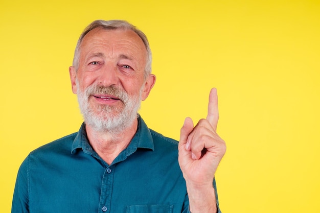 Ritratto ravvicinato di un vecchio anziano felice che guarda la fotocamera, indossando una camicia di cotone verde con un colletto che punta il dito verso l'alto per copiare spase in studio su sfondo giallo.