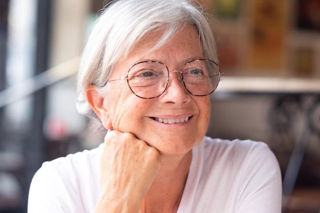 Ritratto ravvicinato di una bella donna caucasica anziana con capelli corti bianchi e occhiali mentre guarda pensierosa lontano mentre è seduta in una caffetteria