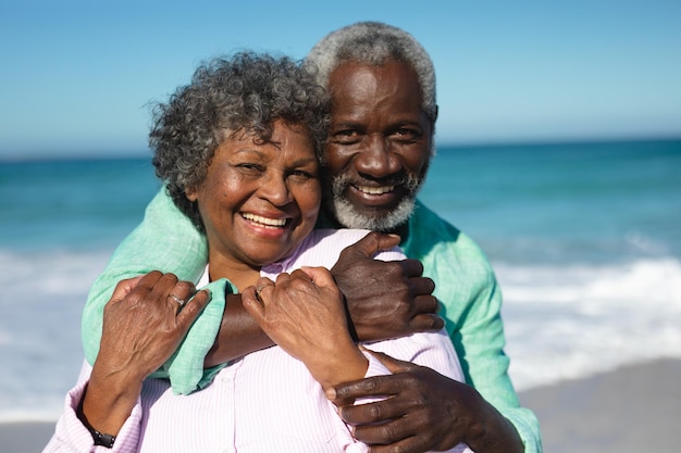Ritratto ravvicinato di una coppia afroamericana anziana in piedi sulla spiaggia con cielo blu e mare sullo sfondo, abbracciando e sorridendo alla telecamera