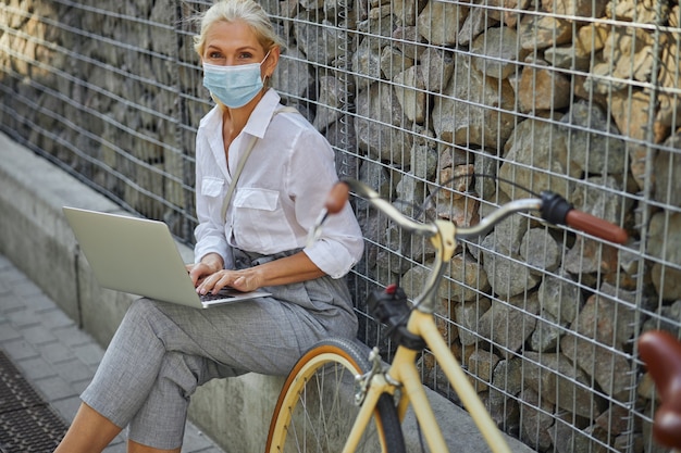 Ritratto ravvicinato di una donna sorridente che manda SMS su un laptop grigio mentre è seduto di fronte al muro di pietra in città