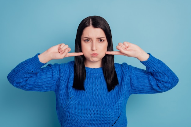 Ritratto ravvicinato di una ragazza spensierata che scherza allegra giocosa, eccitata, che tiene l'aria nelle guance isolate su sfondo blu